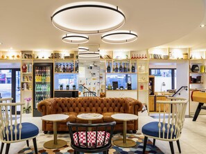 A cozy lounge area with a brown leather sofa, two round tables, and chairs. There are shelves with books and decorative items, and a refrigerator with beverages.