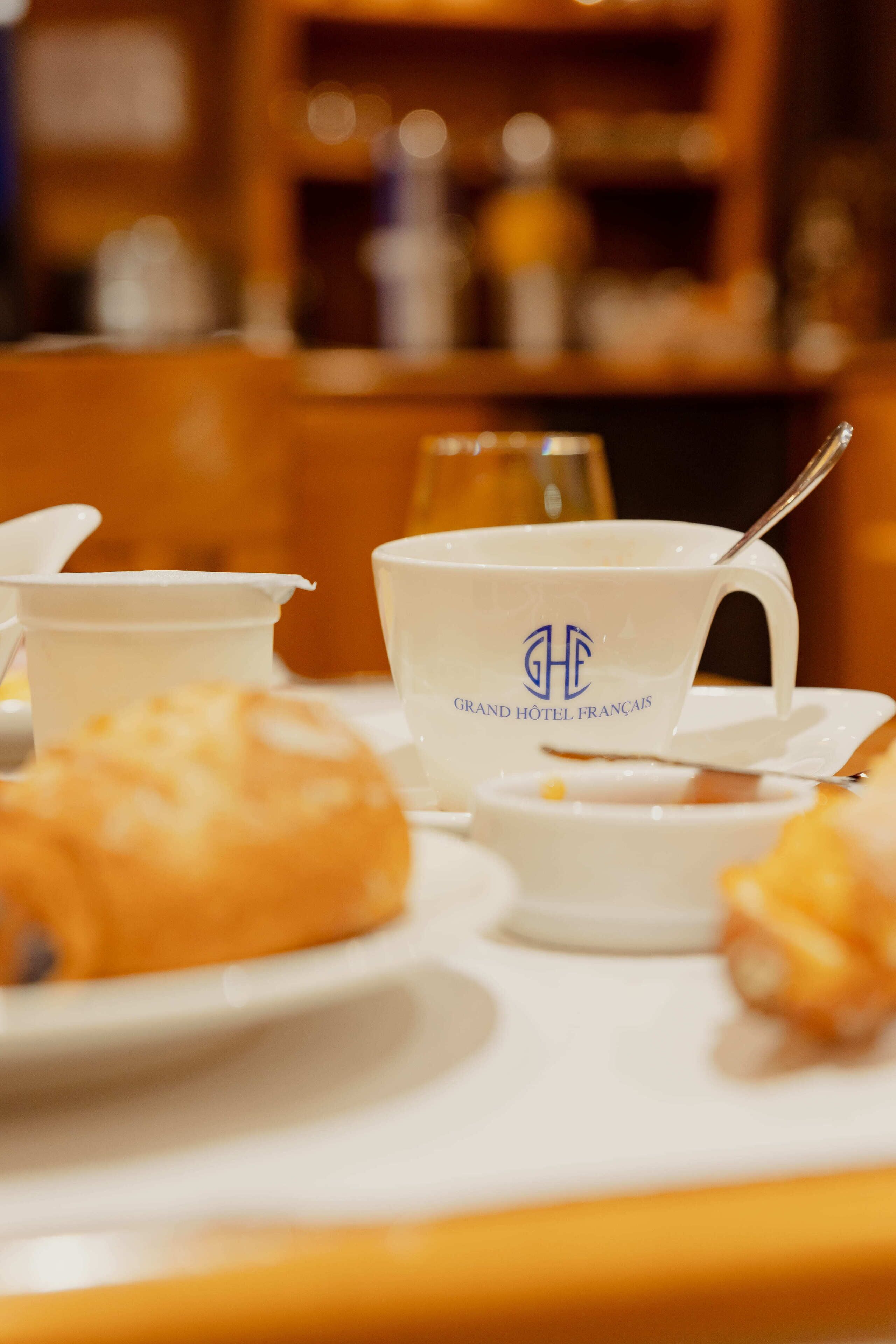 A breakfast setting with a cup labeled "Grand Hôtel Français," a plate with pastries, and a glass of orange juice.