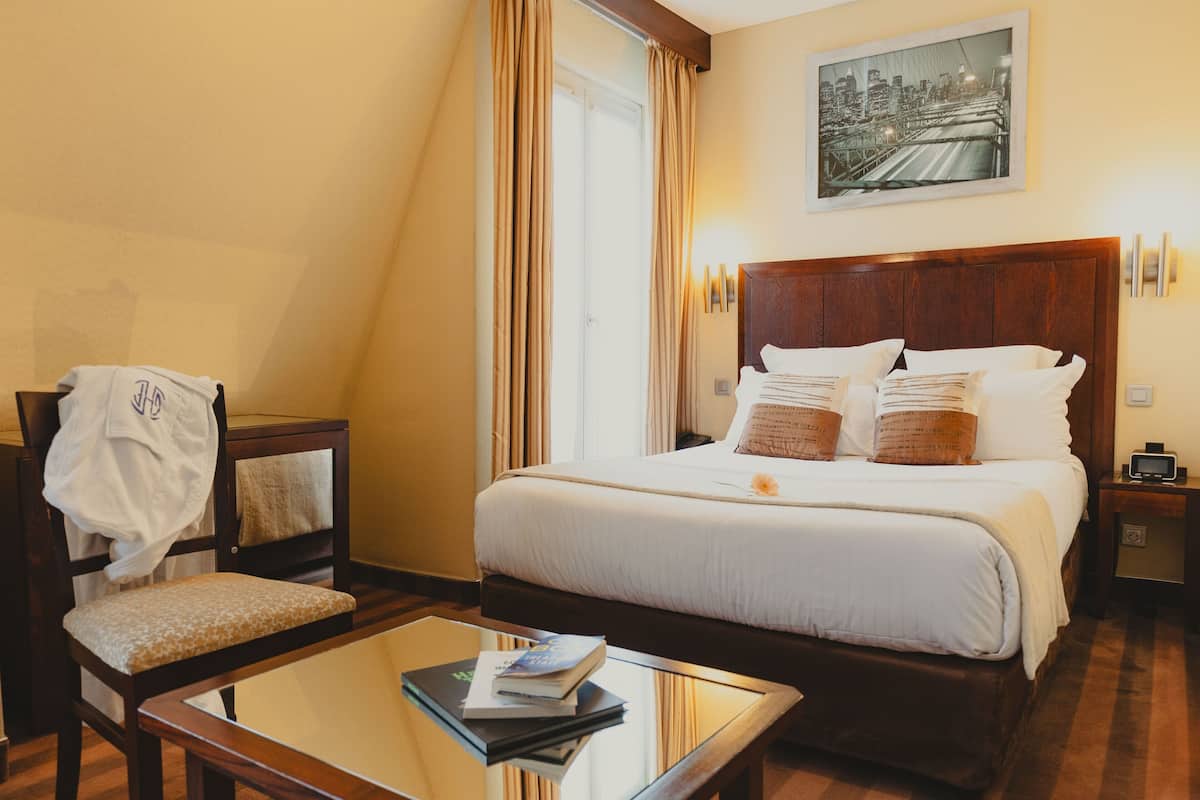 A hotel room with a large bed, a chair, a small table, and a cityscape painting on the wall.