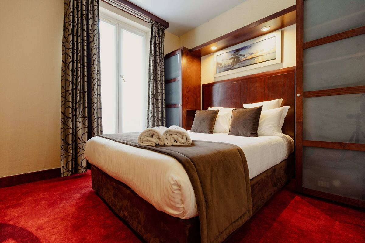 A hotel room with a large bed, a wooden headboard, a red carpet, and a window with curtains.