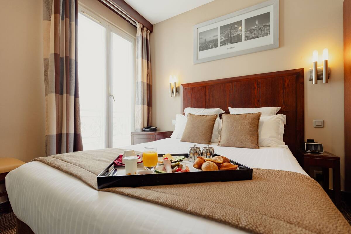 A hotel room with a neatly made bed, breakfast tray with croissants, juice, and a coffee pot.