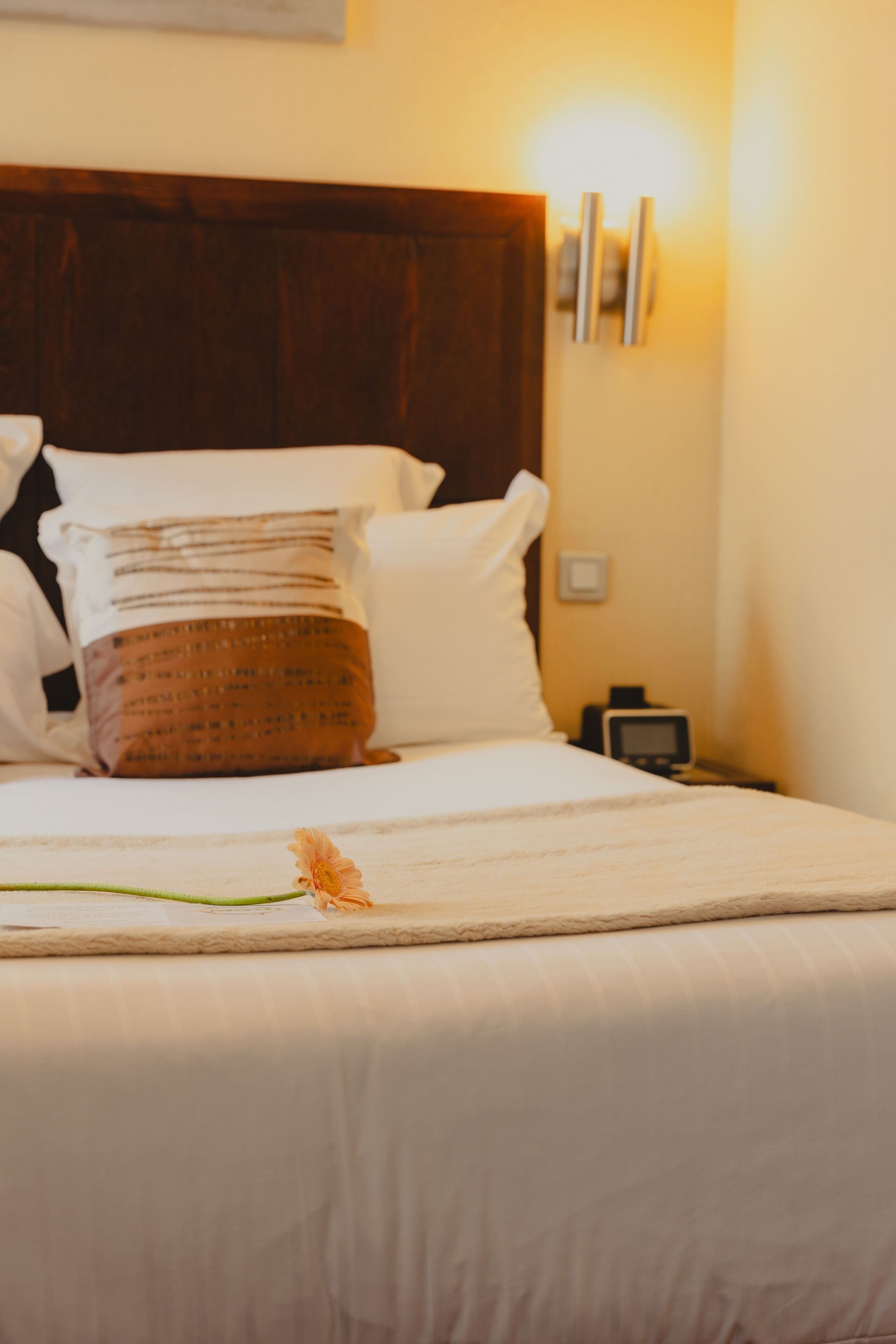 A neatly made bed with a patterned pillow, a bedside table with a clock, and a wall-mounted light fixture.