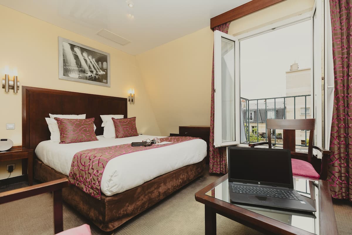 A hotel room with a large bed, a laptop on a desk, a window with curtains, and a framed picture on the wall.