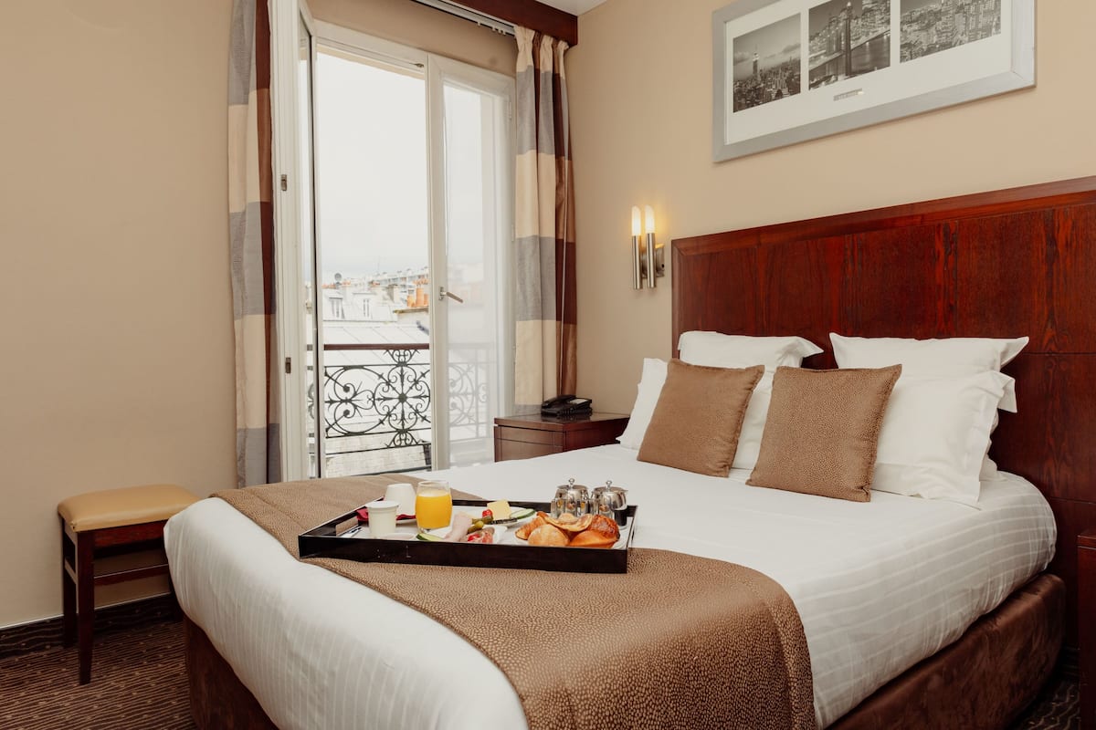 A hotel room with a bed, a tray with breakfast items, a nightstand, a chair, and a balcony with a railing.