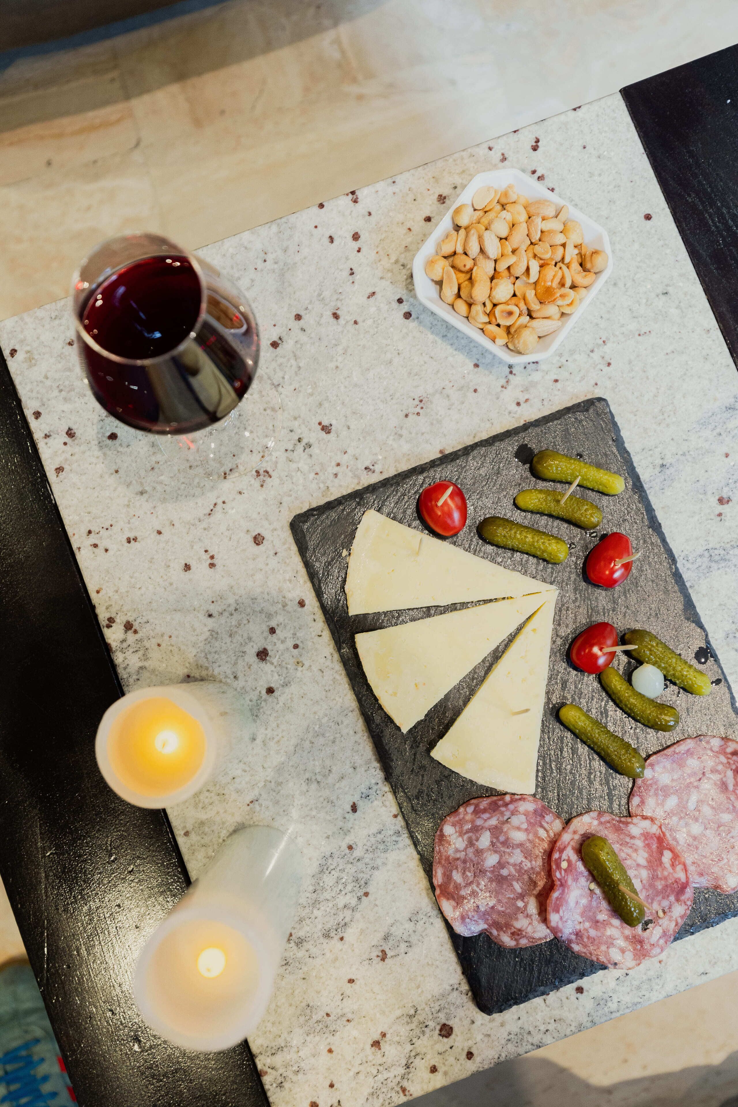 A charcuterie board with cheese, salami, pickles, and nuts on a slate board, accompanied by a glass of red wine and lit candles.