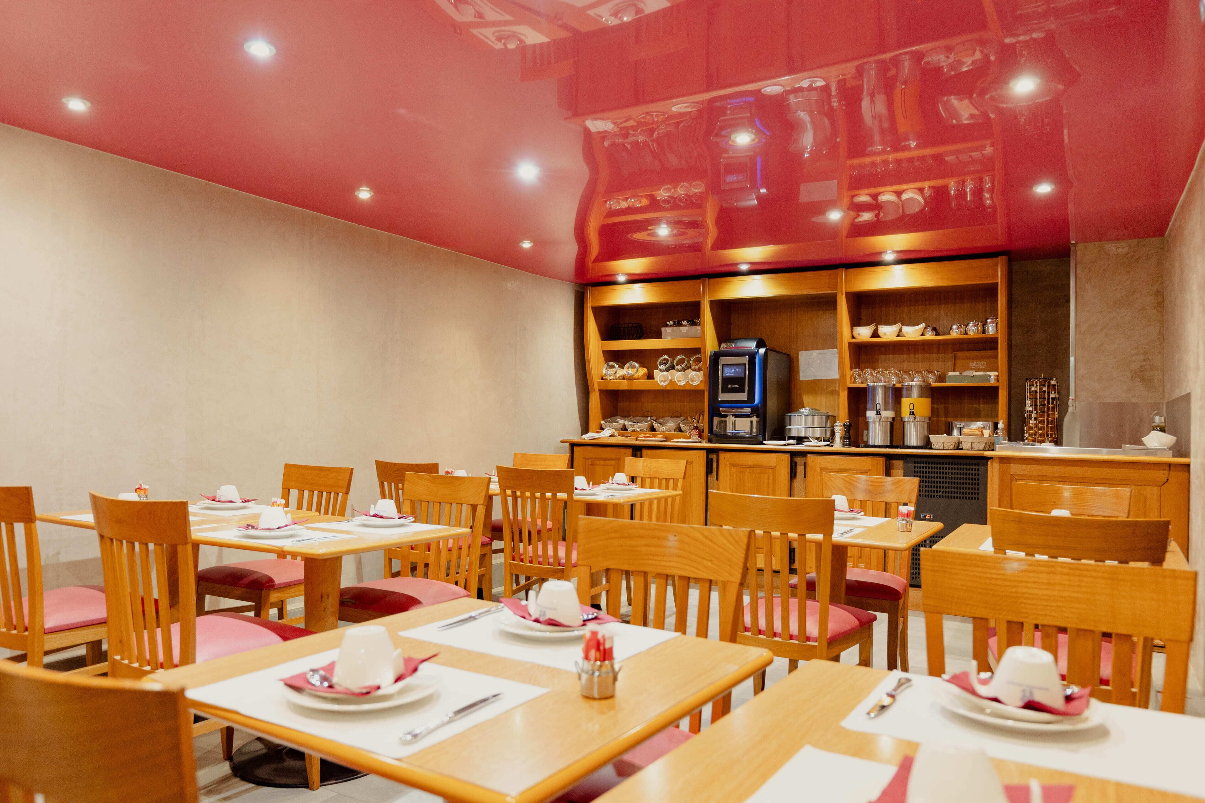Un restaurant avec des tables et des chaises en bois, un espace cuisine et des étagères remplies d’assiettes et de verres.