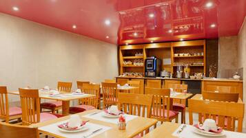 Un restaurant avec des tables et des chaises en bois, un espace cuisine et des étagères remplies d’assiettes et de verres.