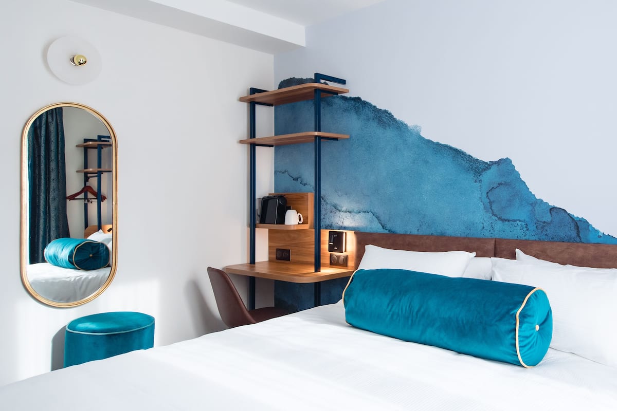 Una habitación de hotel con una cama, un escritorio con silla, un espejo y una pared con un diseño abstracto en azul.