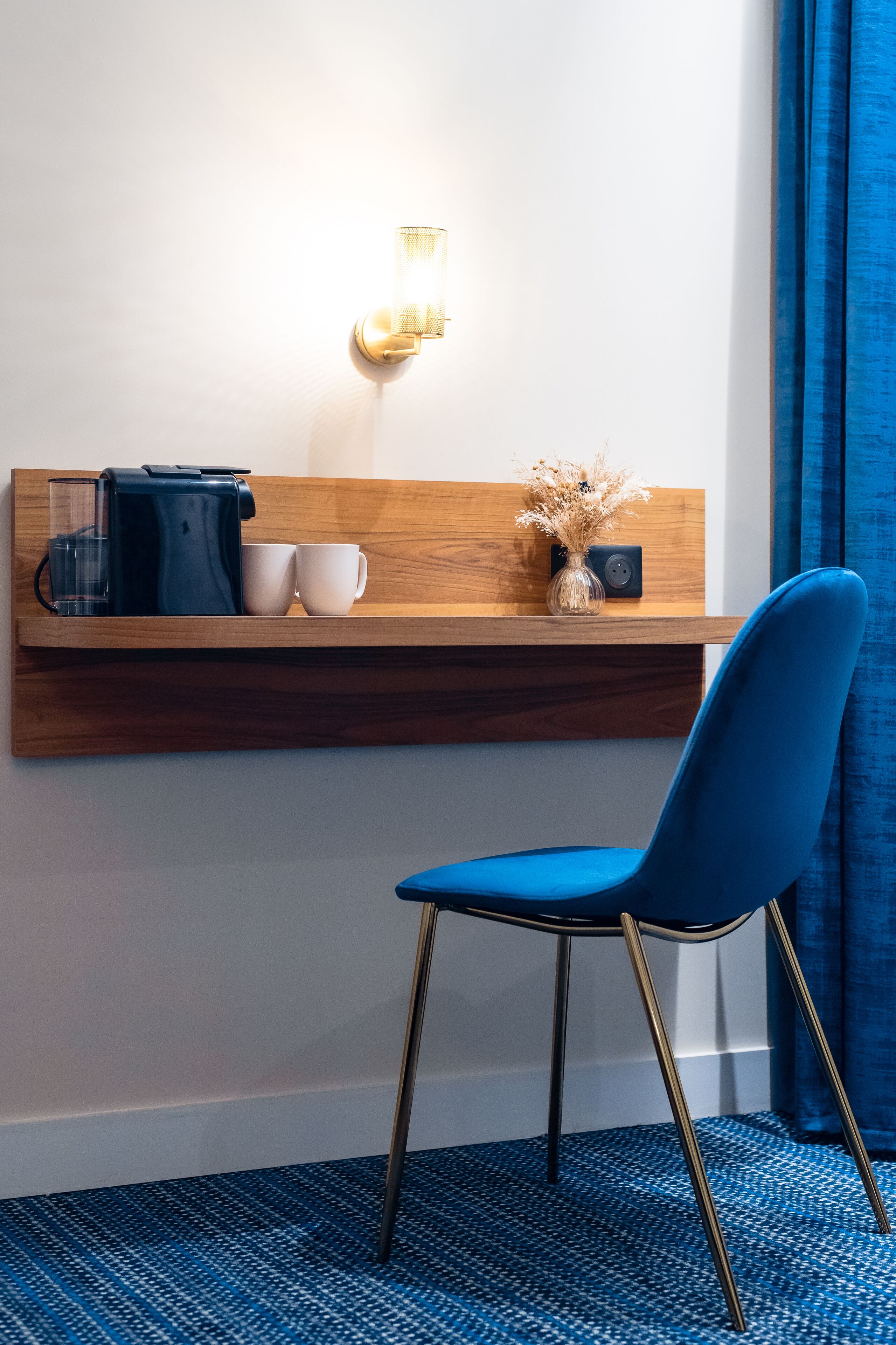 Habitación de hotel con una estantería de madera que sostiene una cafetera, dos tazas y un jarrón con flores secas. Una silla azul está ubicada frente a la estantería.