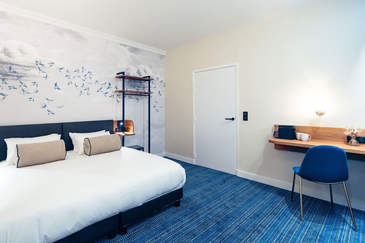 Habitación de hotel con cama, escritorio, silla, puerta y pared decorada con un mural de aves.