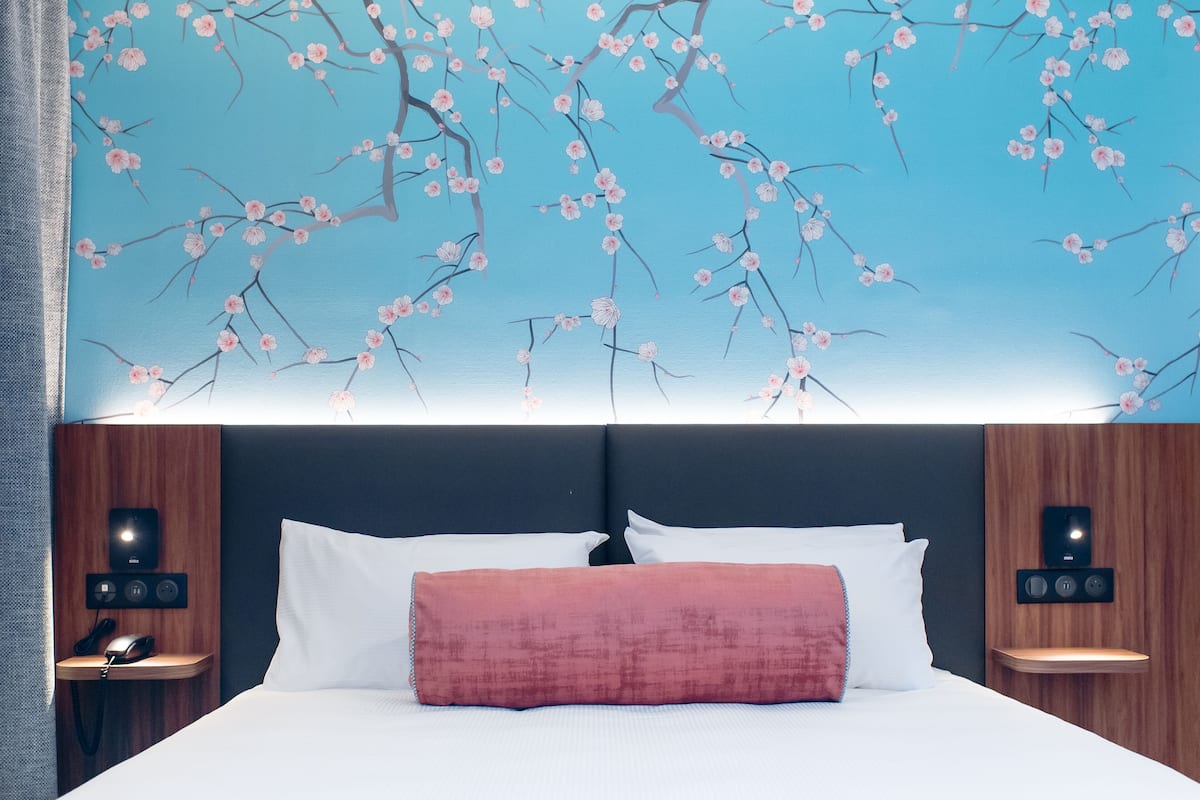 Habitación de hotel con una cama grande, un cojín rojo y un mural de cerezos en la pared. Hay dos mesitas de noche, con lámparas y un teléfono en una de ellas.