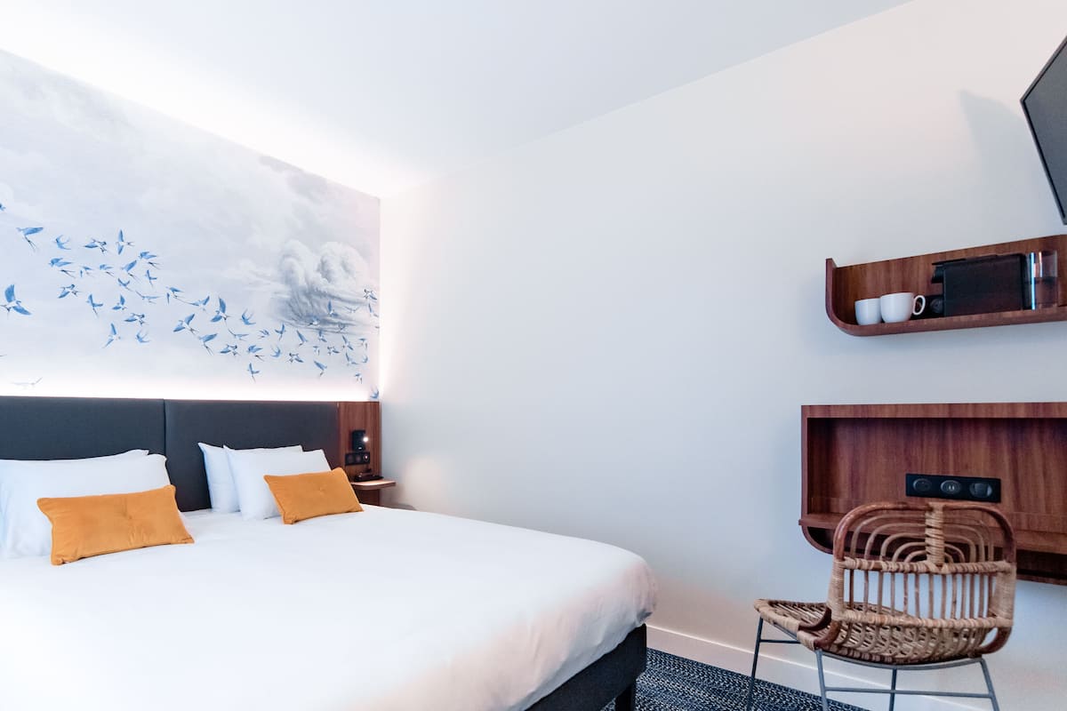 Una habitación de hotel moderna con una cama grande, una silla de madera, un estante en la pared con tazas y un cuadro abstracto de gran tamaño.