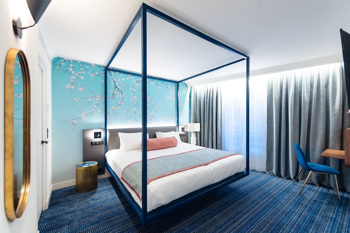Una cama con dosel de color azul, una mesita de noche pequeña con una lámpara, un calefactor en la pared y una ventana con cortinas.
