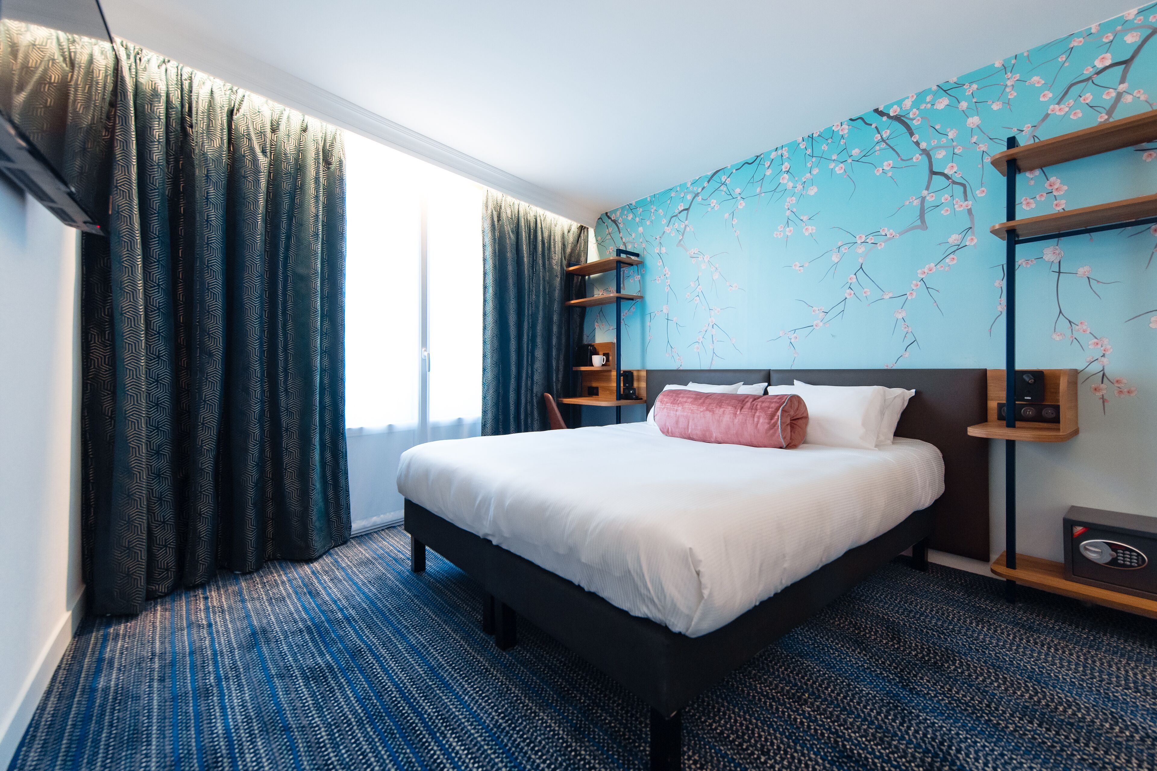 A hotel room with a large bed, a blue patterned carpet, a window with curtains, and a wall with a cherry blossom mural.