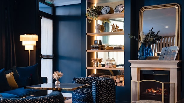 Uma sala de estar moderna com um sofá azul, um banquinho estampado, uma estante, uma lareira e um abajur.