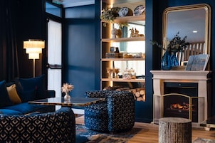 Una sala de estar moderna con un sofá azul, un reposapiés estampado, una estantería, una chimenea y una lámpara.