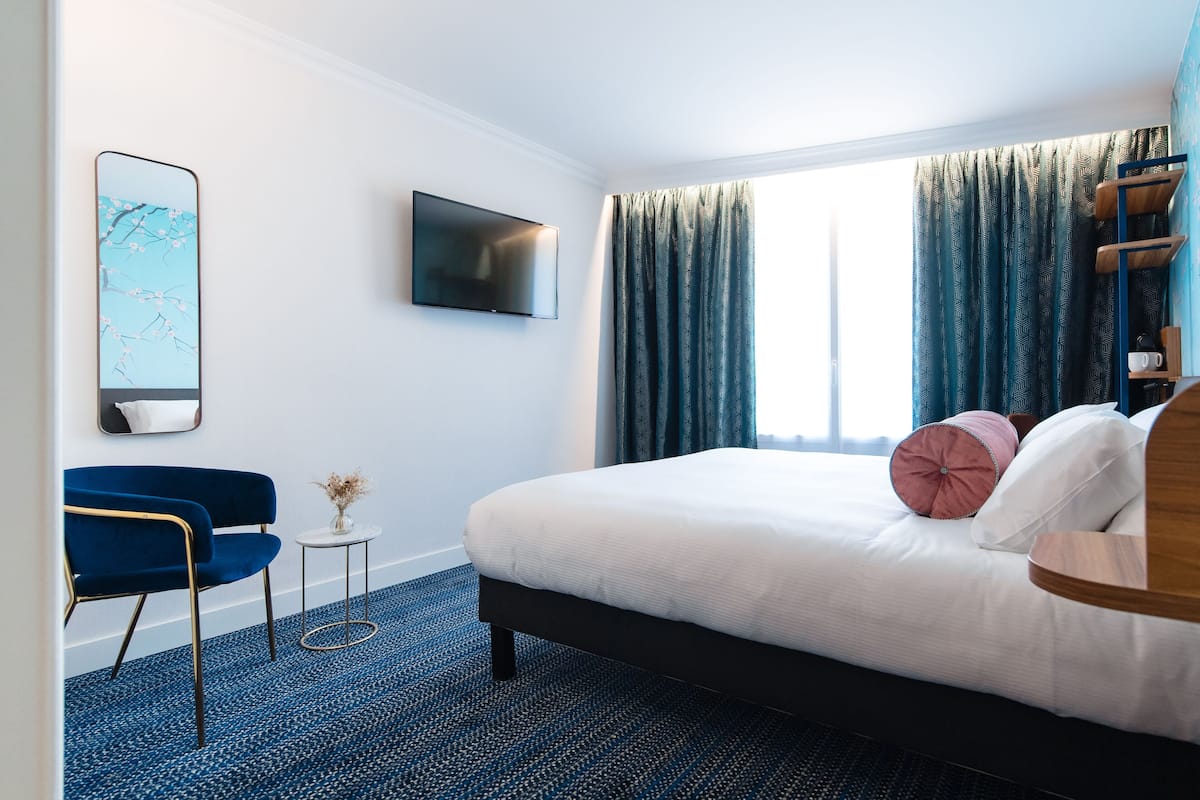 Habitación de hotel con cama, televisor, silla y ventana con cortinas.