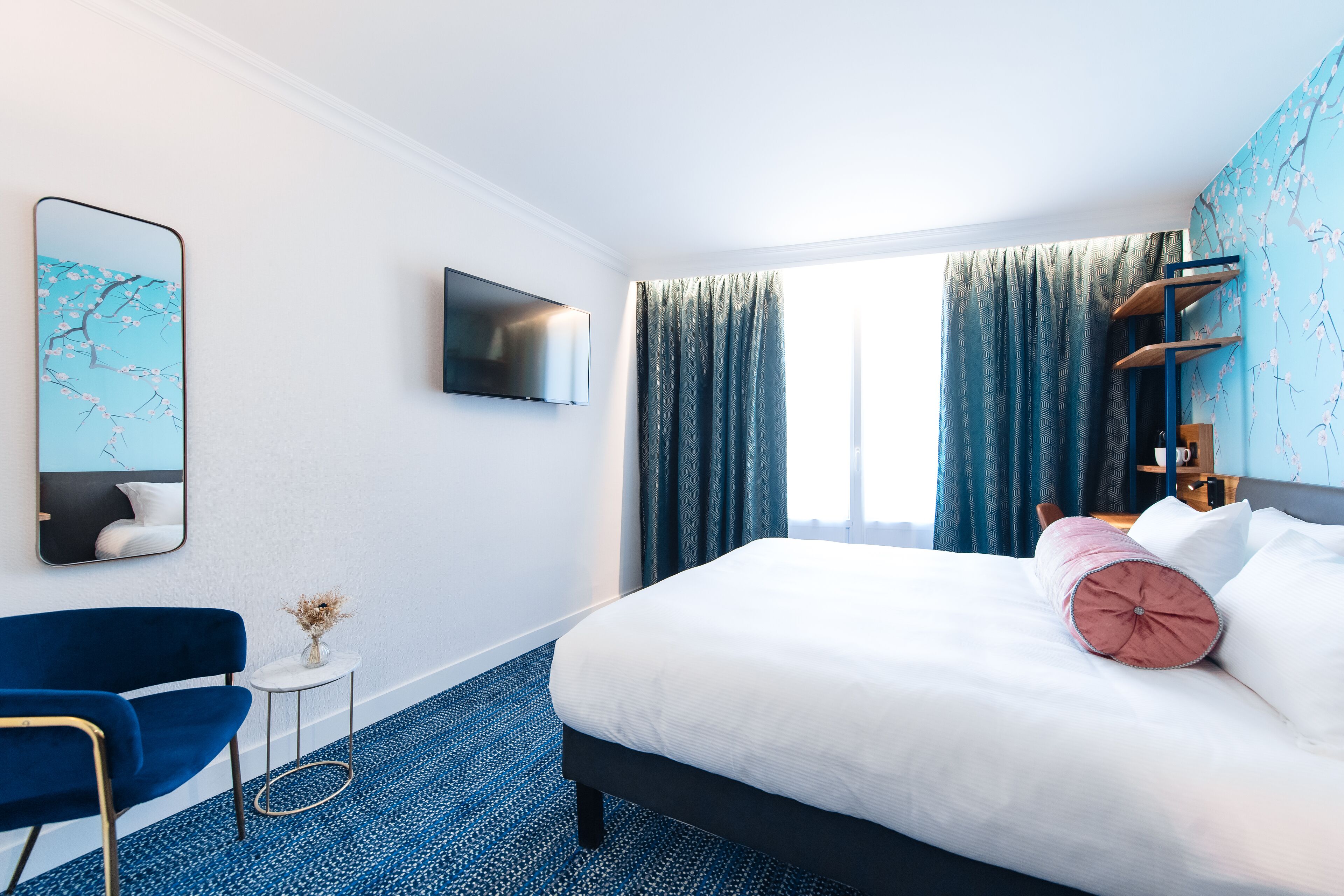 Une chambre d’hôtel avec un lit, une télévision, une chaise, une petite table et une fenêtre avec des rideaux.