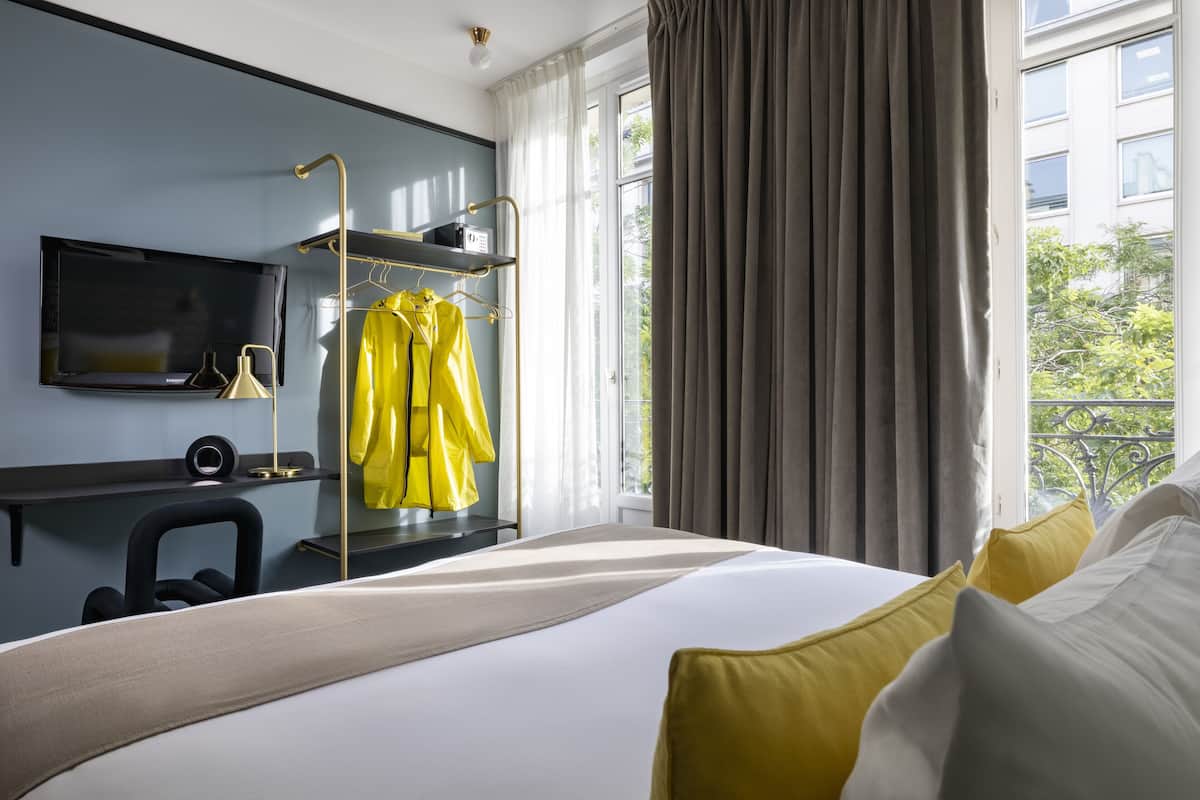 A modern hotel room with a large bed, a TV mounted on the wall, a desk with a chair, and a window with curtains.