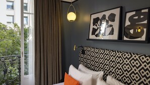 A hotel room with a bed, a patterned headboard, a framed abstract artwork, a hanging lamp, and a view of a building and trees through a window. - Hôtel Palais De Chaillot (Paris)