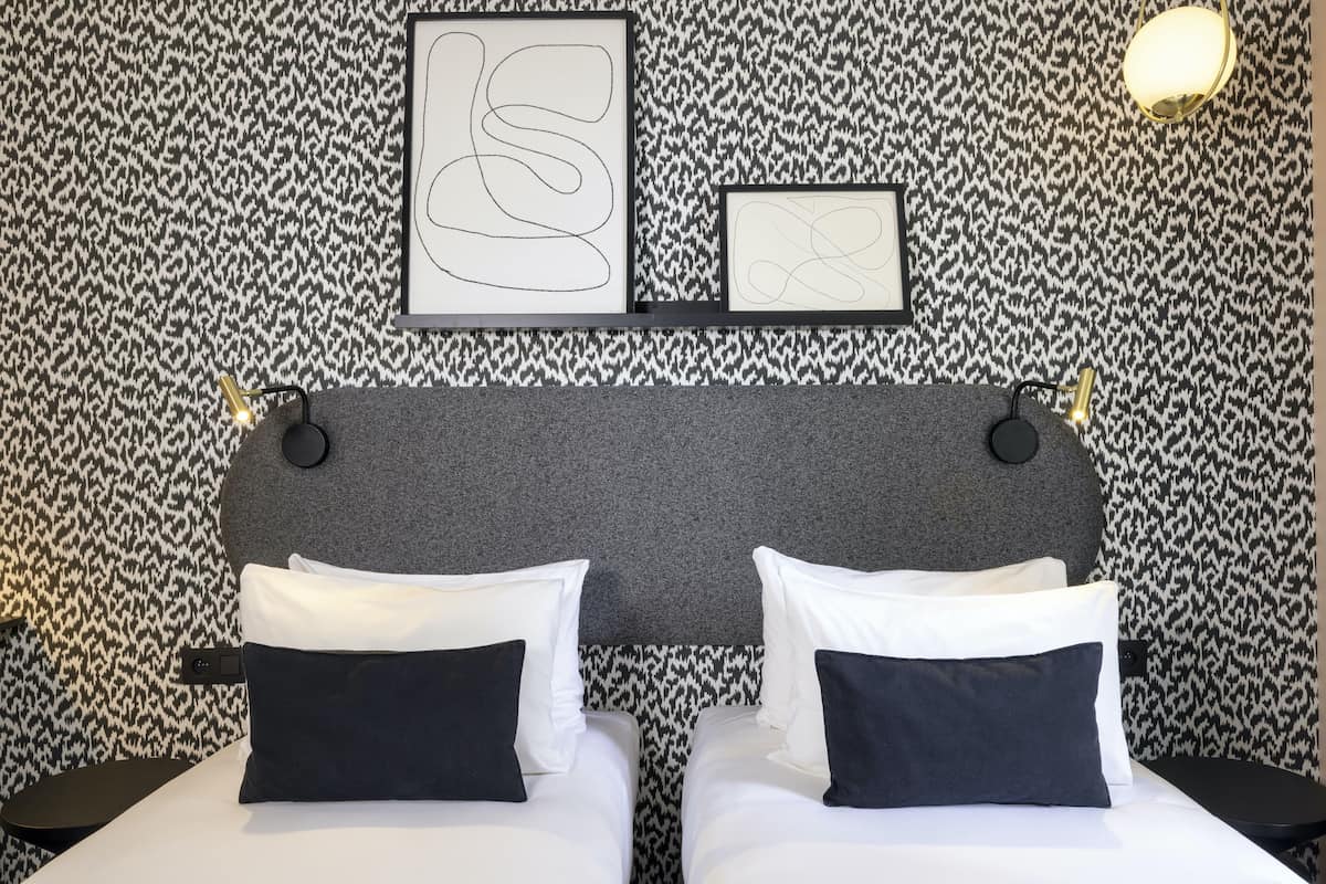 A hotel room with a patterned wallpaper, a grey headboard, two beds with white and navy bedding, and two bedside tables with lamps.