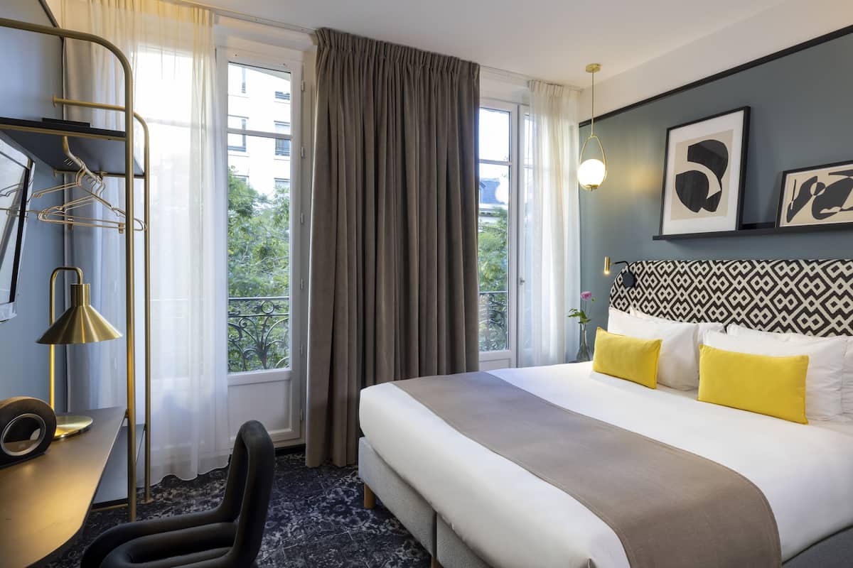 A hotel room with a bed, a desk, a chair, a lamp, and a window with curtains.