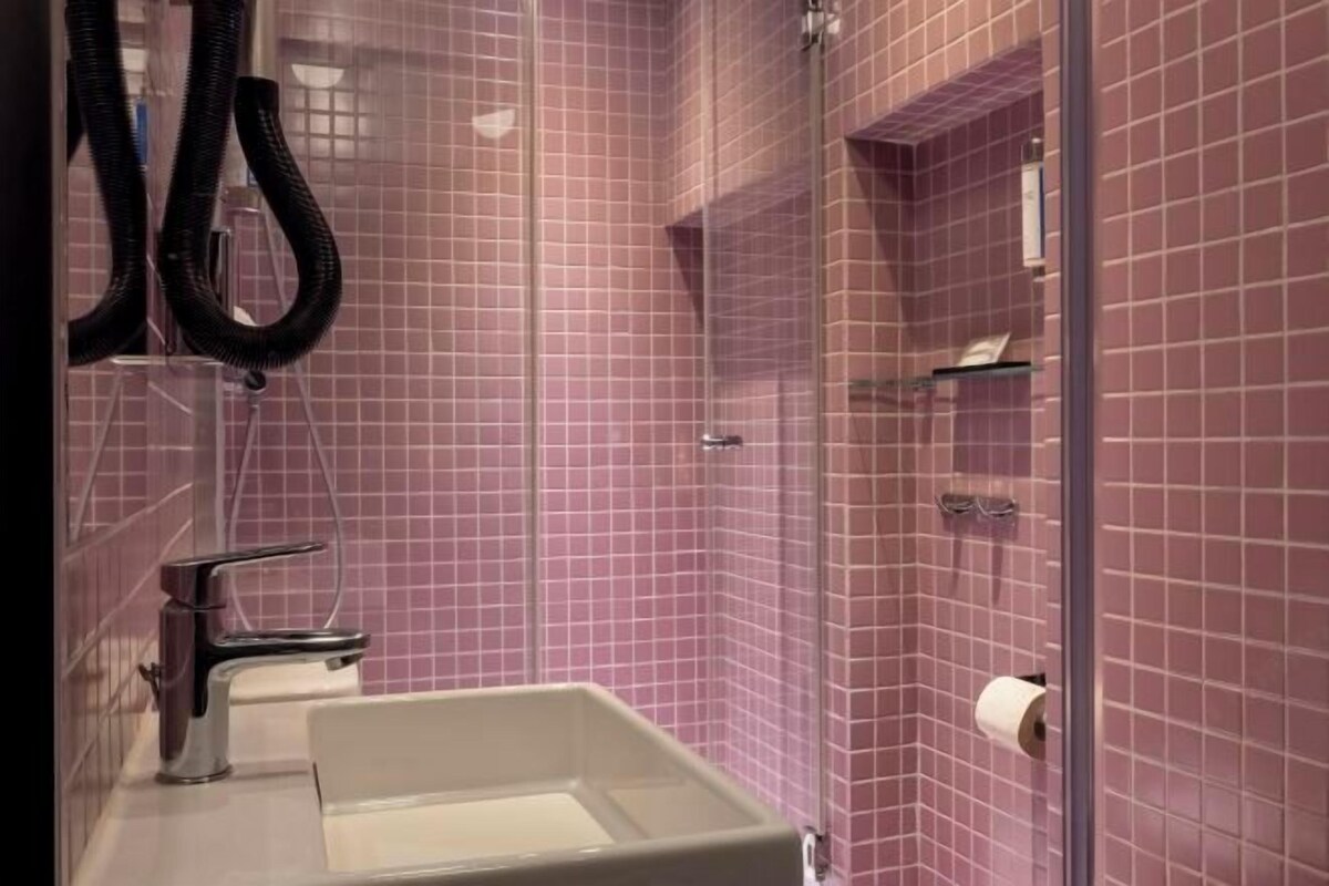 Un bagno con un lavandino bianco, uno specchio e una zona doccia rivestita di piastrelle rosa.