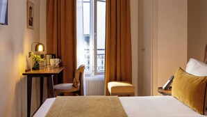 Una camera d'albergo con un letto, una scrivania con una lampada, una sedia, un tavolino con un vaso e una finestra con tende.