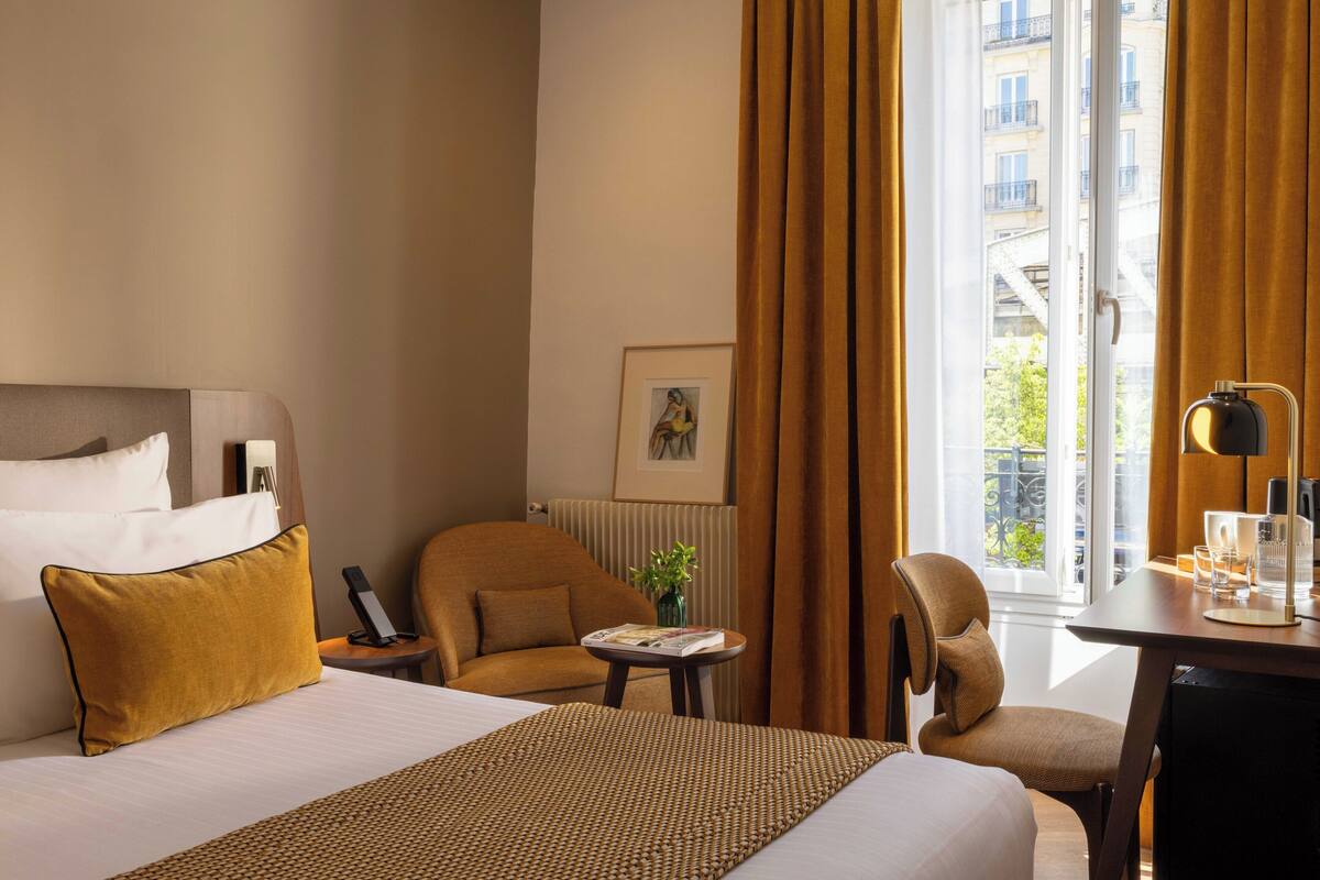 A hotel room with a bed, two armchairs, a small table with a lamp, a framed picture on the wall, and a window with curtains.