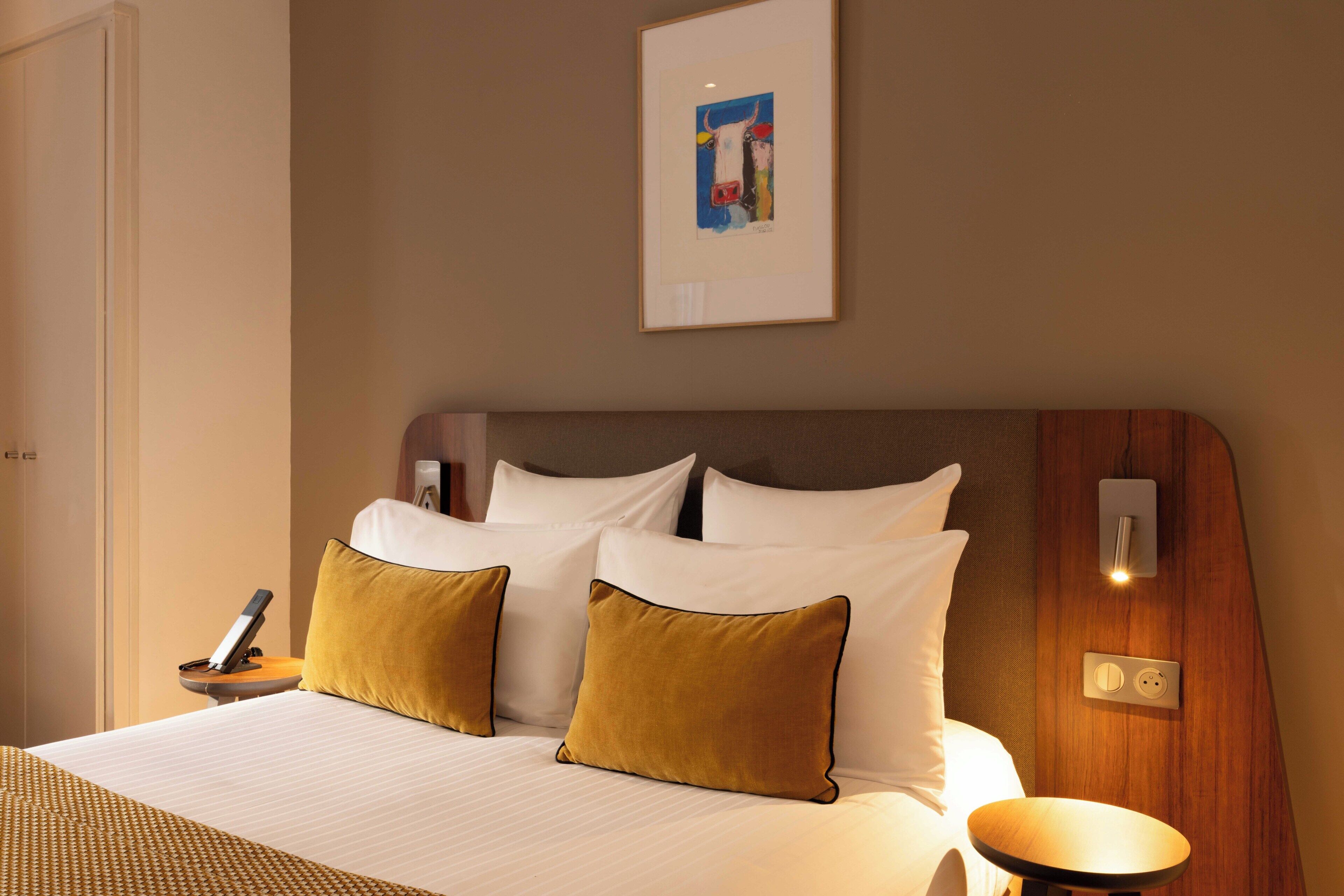 Ein Hotelzimmer mit einem Bett, zwei Kissen, einem Nachttisch mit Telefon, einer Wandleuchte und einem Bild an der Wand.