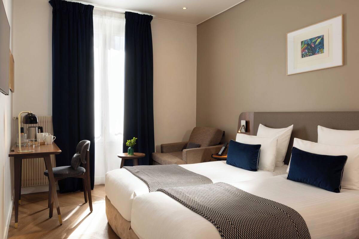 A hotel room with a large bed, a desk with a lamp, a chair, and a small table with a plant.