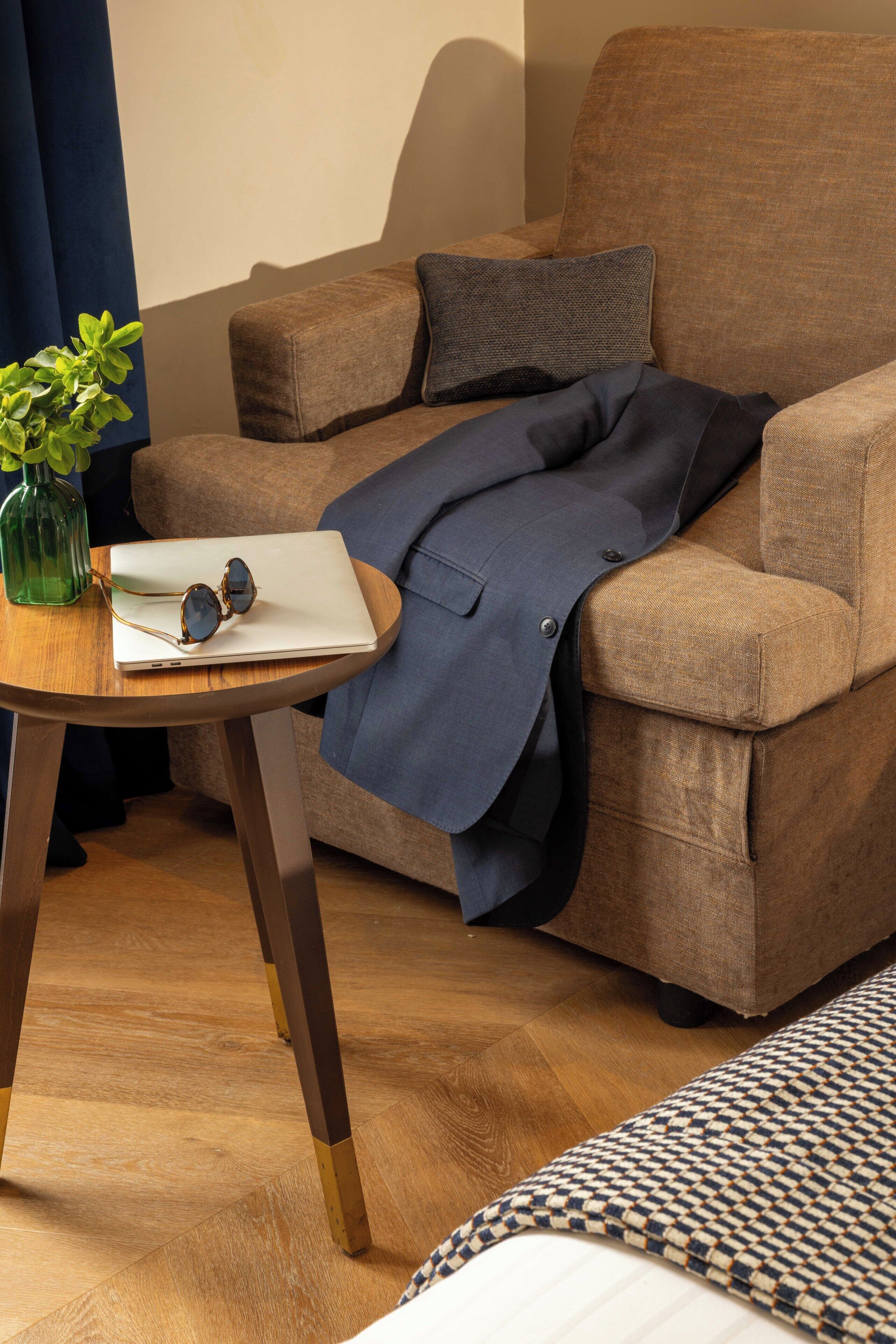 A brown sofa with a blue blanket and a cushion, a wooden table with a plant and a pair of sunglasses, and a patterned rug.