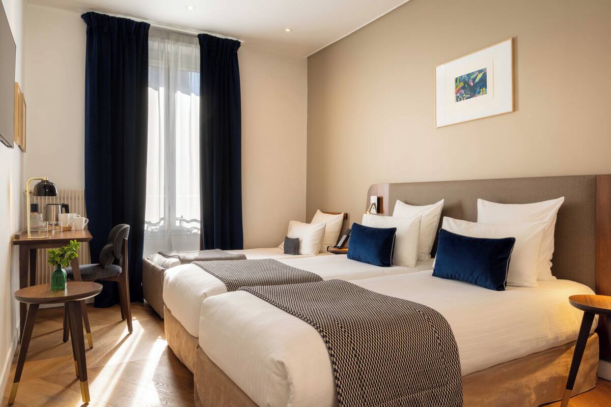A hotel room with two beds, a desk with a chair, a small table with a plant, and a window with curtains.