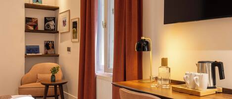 Ett hotellrum med en säng, ett skrivbord med en lampa, en stol, en kaffebryggare och en hylla med böcker och bilder.