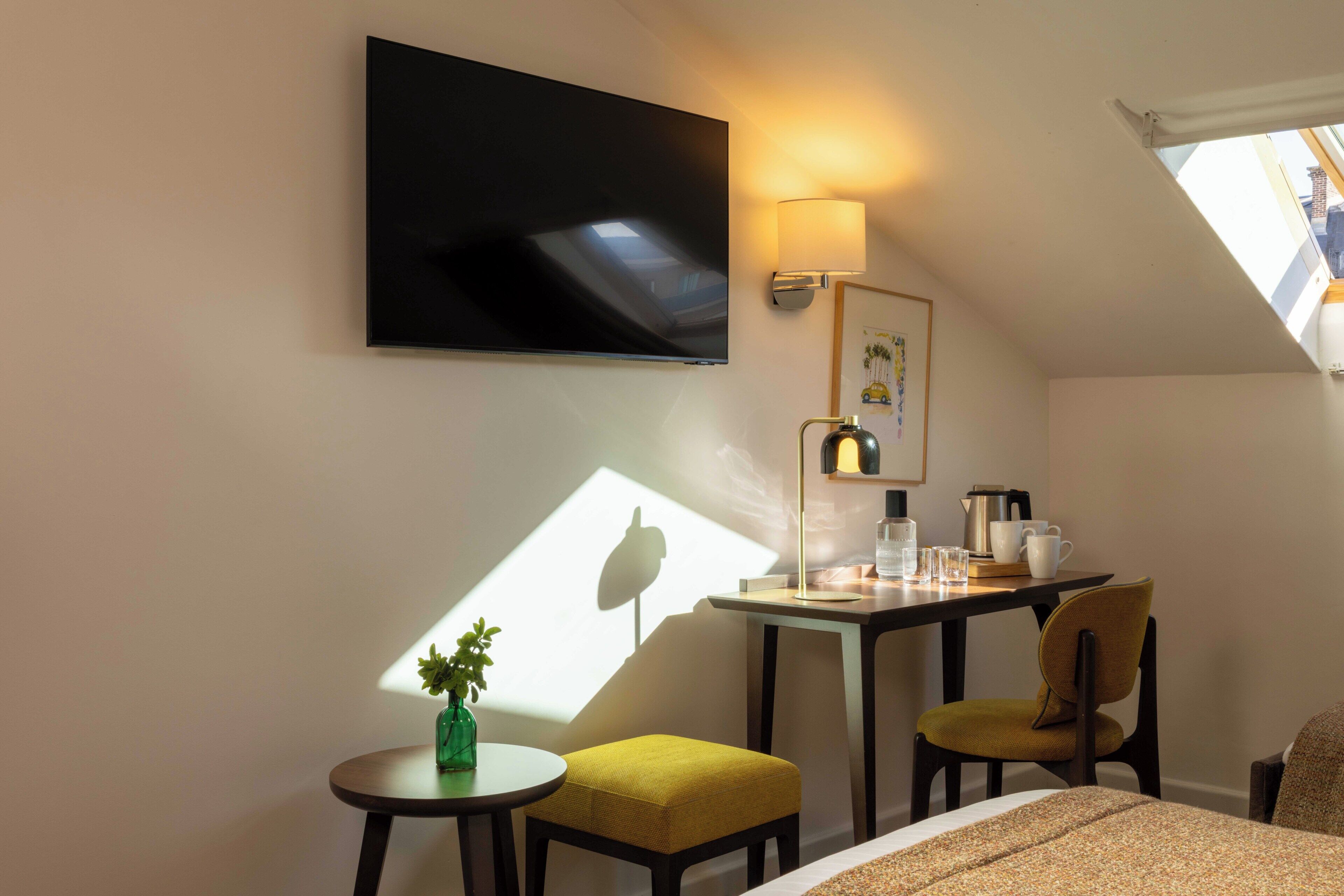 A hotel room with a bed, a desk with a lamp, a chair, a small table with a plant, and a TV mounted on the wall.