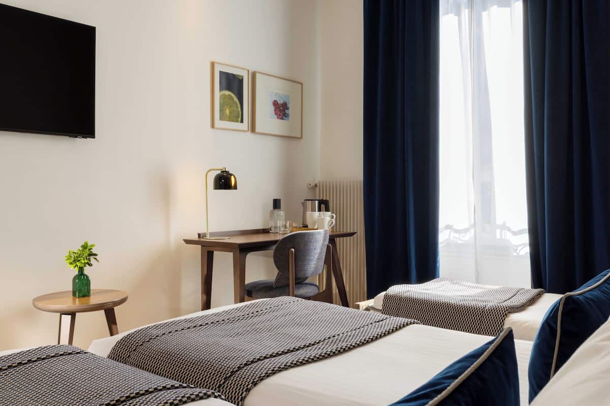 A hotel room with two beds, a desk with a chair, a small table with a plant, and artwork on the wall.