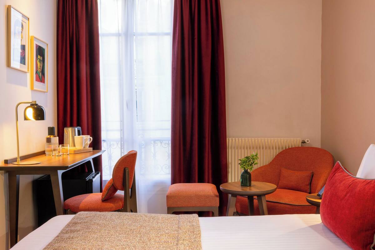 A hotel room with a bed, a desk with a lamp, a chair, a small table with a plant, and a window with curtains.