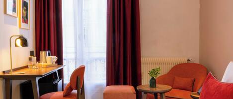 Ein Hotelzimmer mit Bett, Schreibtisch mit Lampe, Stuhl, kleinem Tisch mit Pflanze und Fenster mit Vorhängen.
