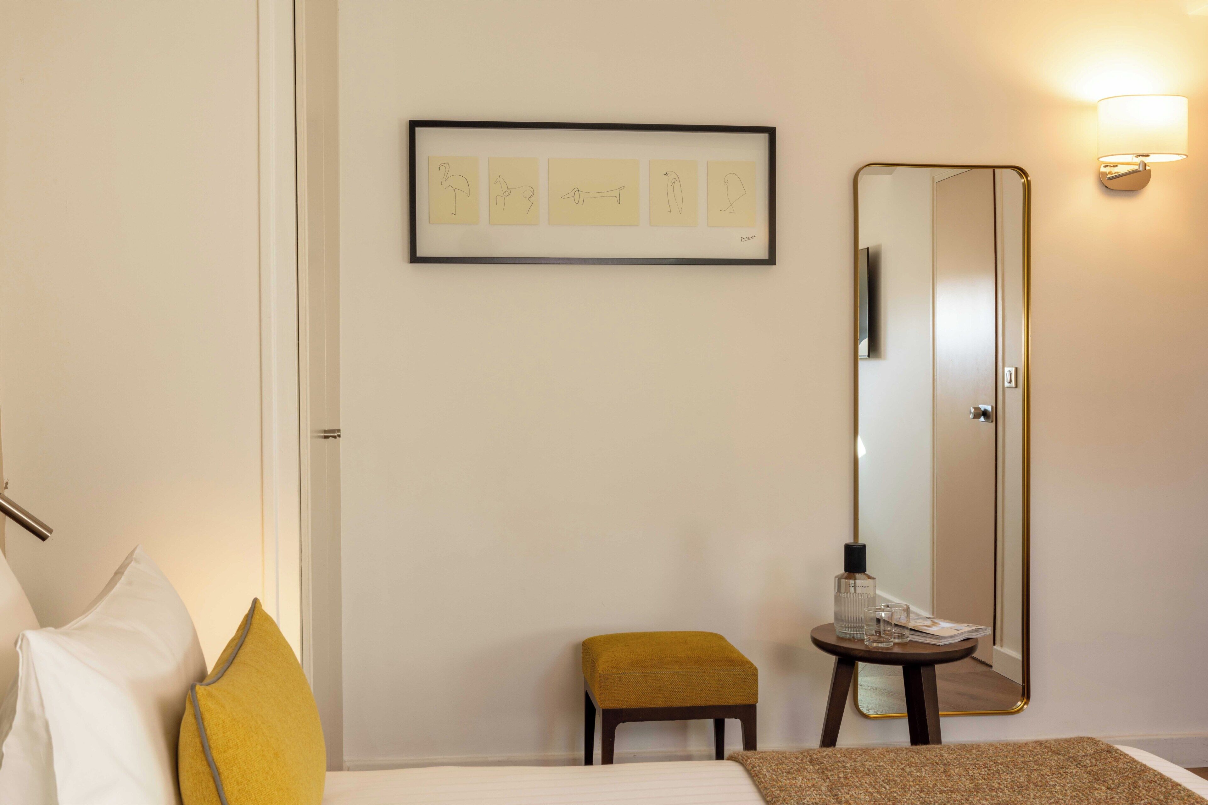 A hotel room with a bed, a small stool, a framed artwork, a mirror, and a wall-mounted lamp.