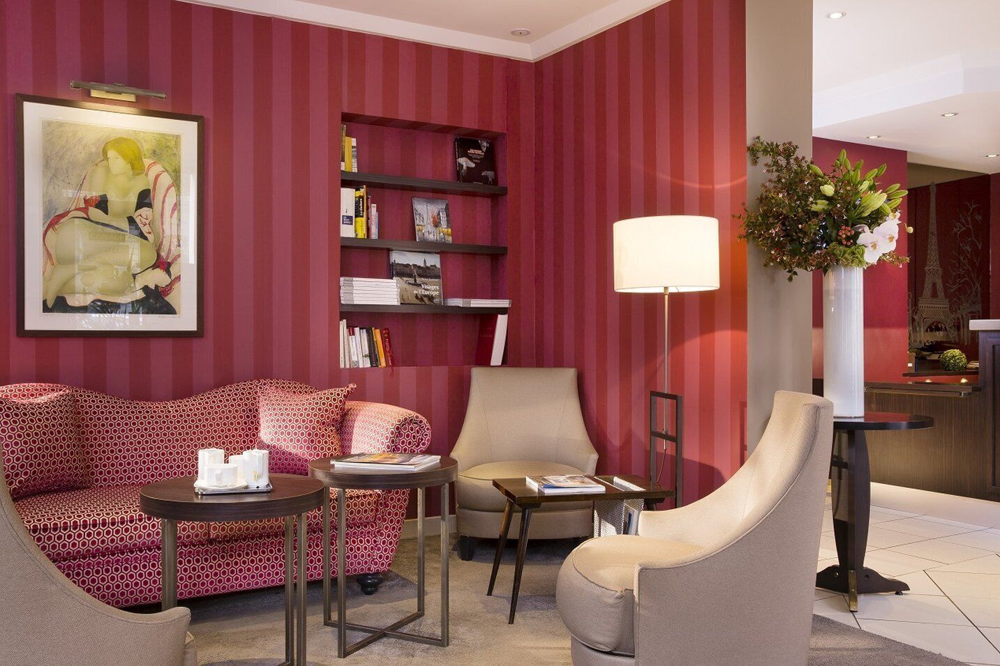 A hotel room with a red-striped wall, a sofa, a chair, a small table, a bookshelf, and a floor lamp.