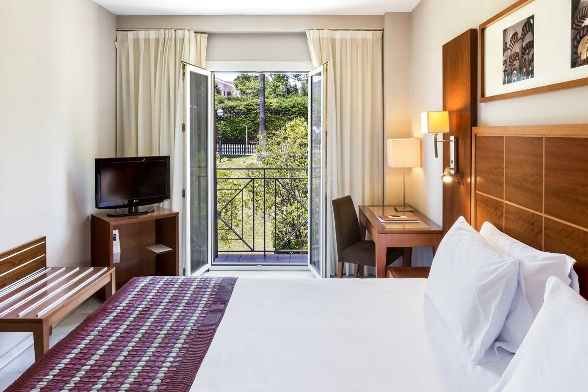 A hotel room with a bed, a desk, a TV, and a balcony with a view of greenery.