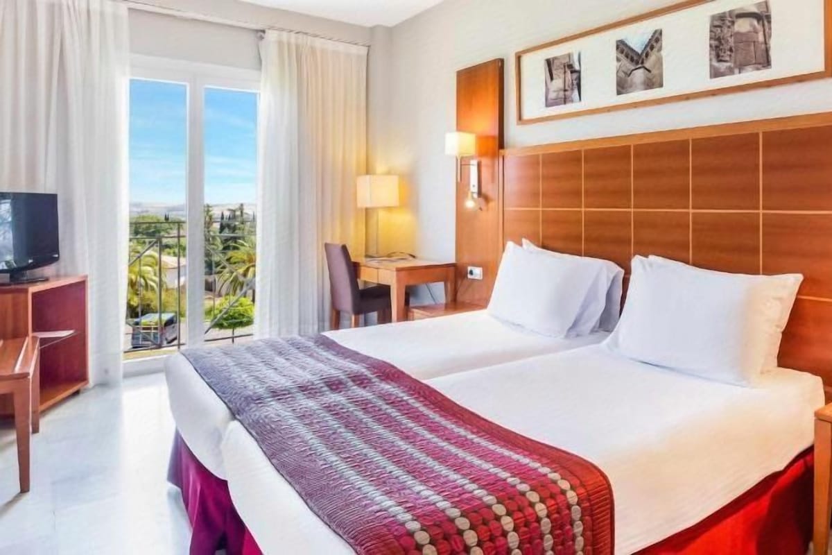 A hotel room with a large bed, a desk, a chair, a TV, and a balcony with a view.