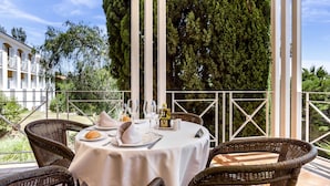 Un área de comedor al aire libre con sillas de mimbre y una mesa redonda para dos personas, rodeada por una barandilla blanca y follaje.