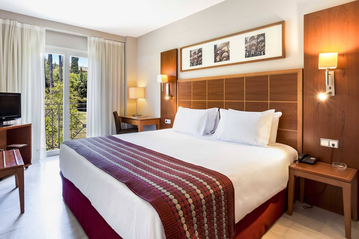 A hotel room with a large bed, a desk, a chair, a TV, and a balcony with curtains.