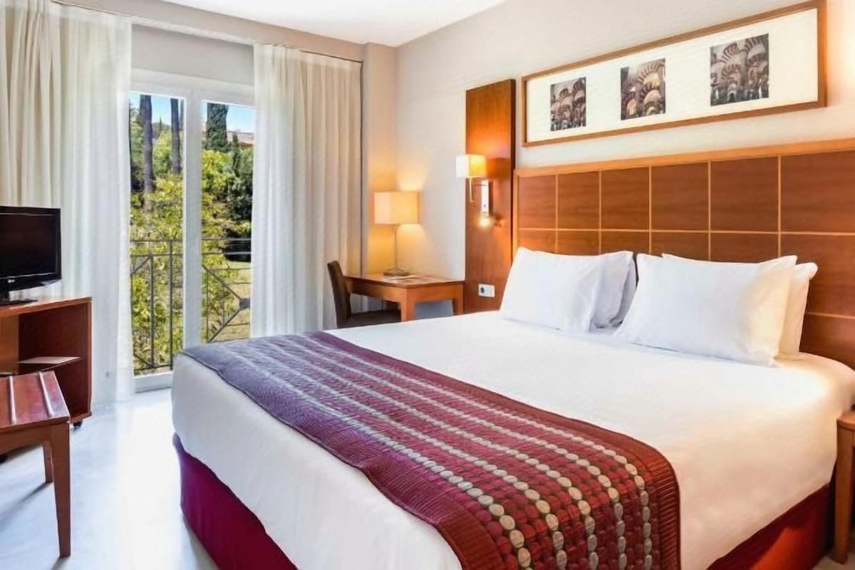 A hotel room with a large bed, a television, a desk, and a balcony with curtains.