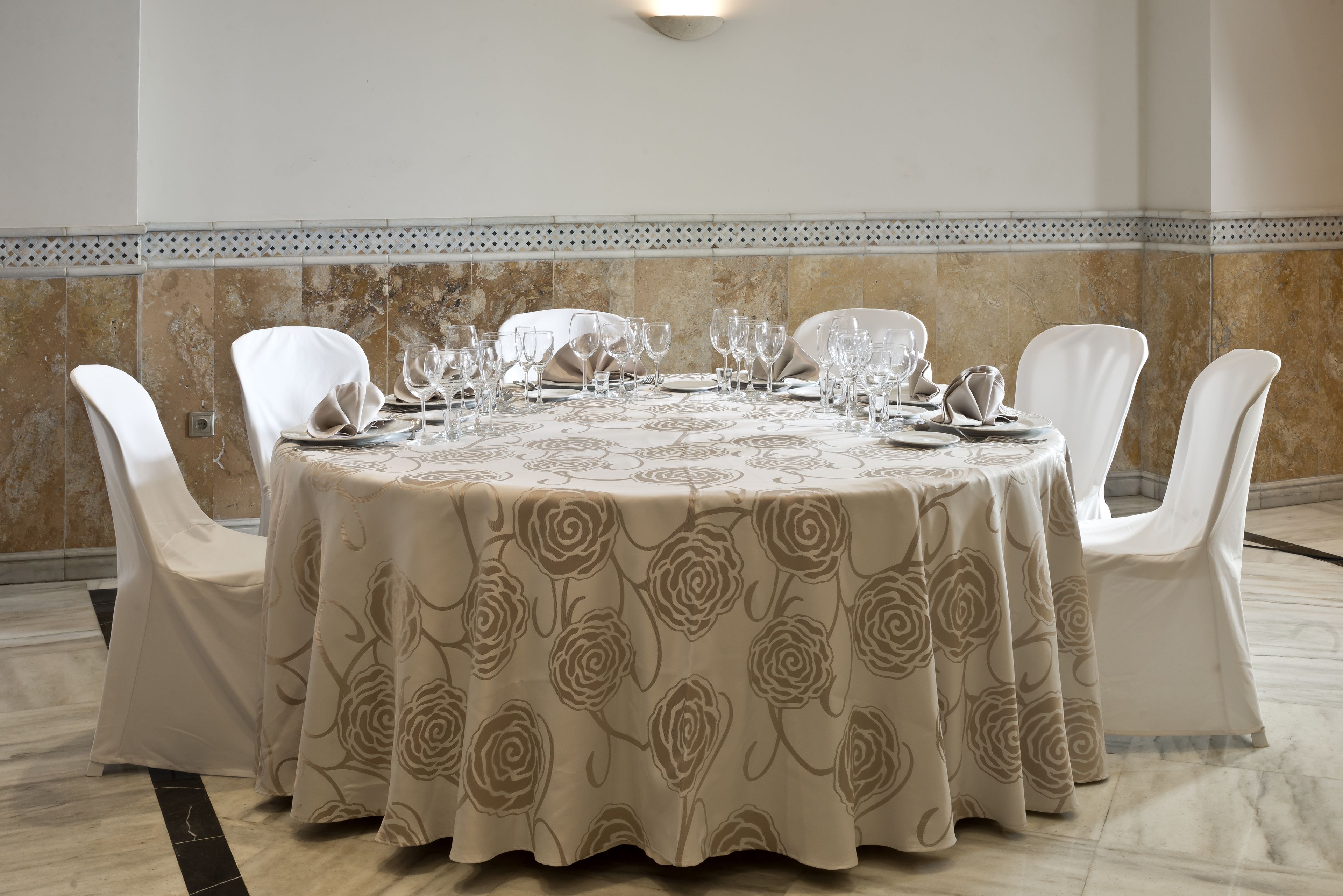 Une table ronde recouverte d’une nappe à motifs floraux, entourée de chaises blanches, dans une pièce dont un mur est recouvert de carreaux et orné d’une bordure décorative.