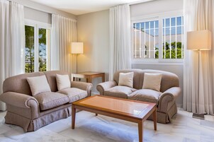 En moderne stue med to beige sofaer, et træbord og et lille sofabord. Værelset har store vinduer med hvide gardiner, der giver udsigt til grønne omgivelser udenfor.