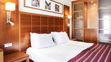 Una habitación de hotel con una cama grande, cabecera de madera, dos mesitas de noche, un armario con puertas de vidrio y cuadros en la pared.