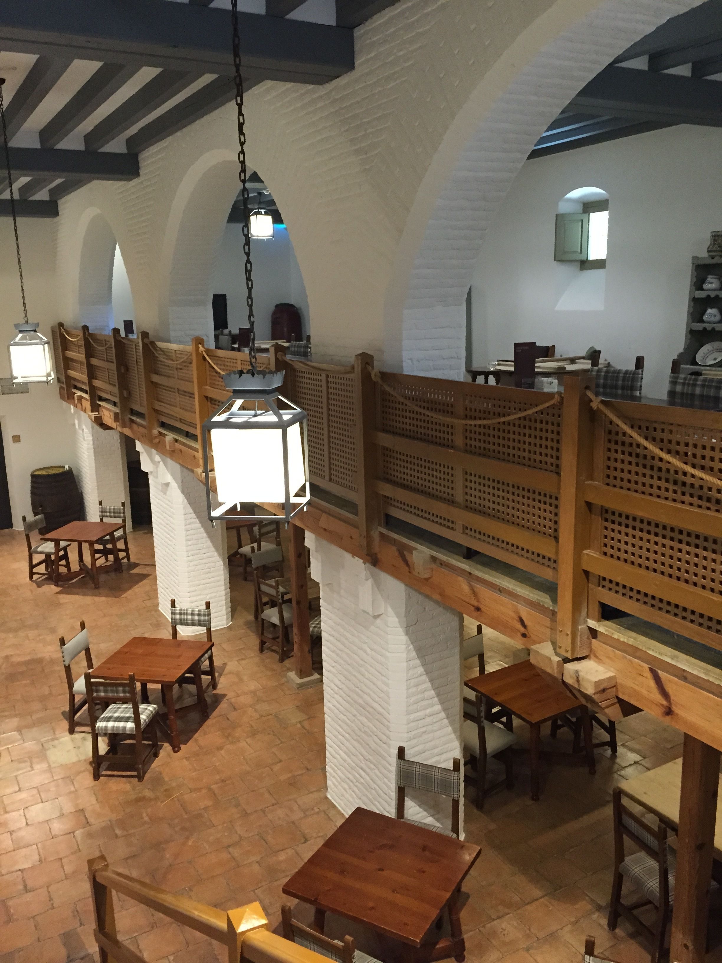 Un restaurante con techos altos de madera, paredes de ladrillo blanco y vigas a la vista. Mesas y sillas dispuestas para comer.