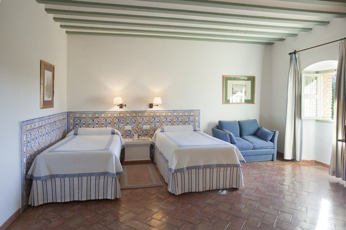 A hotel room with two beds, a blue sofa, a tiled headboard, and a view of the outdoors.